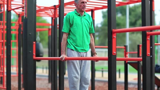 Elderly grey-haired sportsman makes physical exercise on sports ground. Active elderly athlete in green t-shirt trains in open air gym. Aged grey-haired man makes strenuous exercise on parallel bars