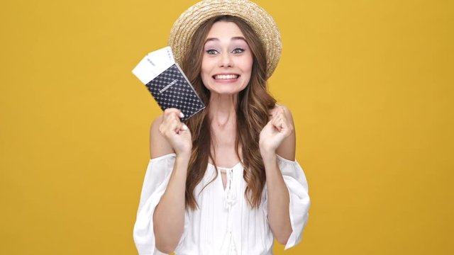 Happy brunette woman in dress and hat holding her pasport with tickets over orange background
