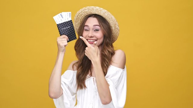 Happy screaming brunette woman in dress and hat holding her pasport with tickets and pointing on it over orange background
