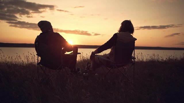 Two young males giving high five each other against beautiful sunset. Camping near a lake, sitting in chairs. Slow motion.