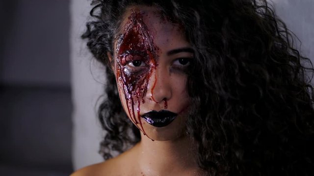Scary portrait of young zombie girl with Halloween blood makeup. Beautiful latin woman with curly hair looking into camera in studio. Slow motion.
