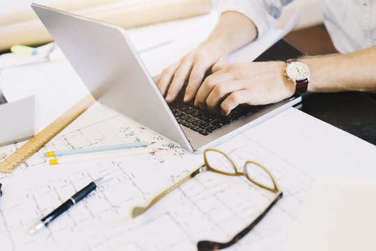 Man using laptop next to construction plan at desk