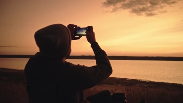 Man taking a picture of beautiful evening sky on mobile phone, sitting in a chair. Slow motion.