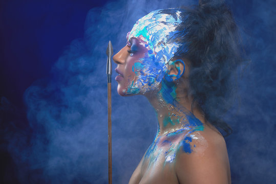 A model with bright fantastic make-up, who keeps an arrow near her face. She stands in a cloud of smoke on the dark blue background and looks like a fantasy charackter.