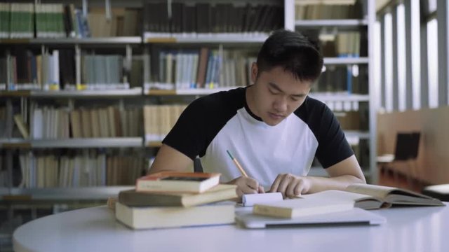 Tracking shot of student studying at desk in library in college. A high school student writes essay in library. Book shelves in background