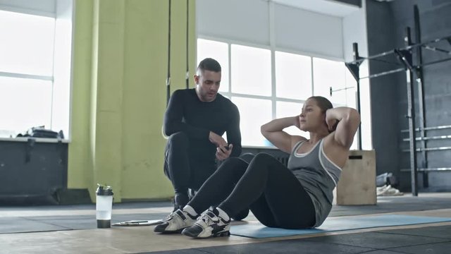Young woman doing crunches in cross training gym while personal coach counting reps with timer