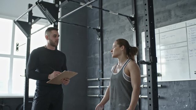 Personal coach holding clipboard and explaining exercise to young muscular woman doing chin-ups on bar in cross training gym