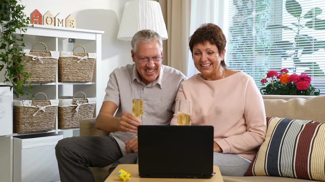 Elderly Couple Connected with Friends using Laptop Video Call Camera and Drinking Champagne Sitting in the Living Room. Slow Motion
