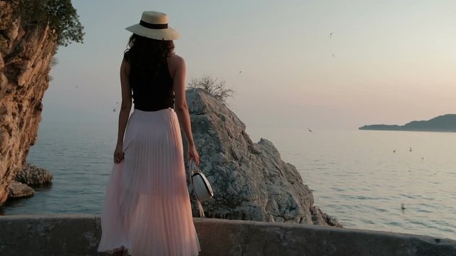 Woman in hat and pleated long skirt is walking along southern shore.
