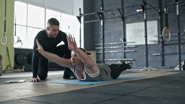 Slowmo of personal coach assisting woman lying on stomach and doing back extension exercise with raising arms and legs in cross training gym