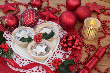 Traditional christmas mince pies on a heart shaped plate with red bauble decorations and  holly with lit candle on oak wood background.