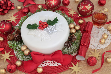 Christmas cake with glass of sherry, snow covered fir, holly, red and gold bauble decorations and foil wrapped chocolates on oak background.