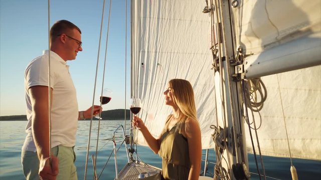 Young couple having a romantic date on the yacht at sunset. Man proposing a toast and they clanging glasses with red wine