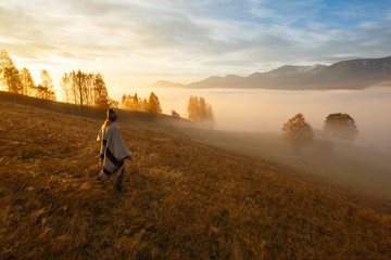 Young woman over the clouds in the valley looking at calm sunrise. Hiker girl wrapping in warm poncho outdoor. Successful woman hiker enjoy the view on mountain top.