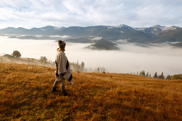 Fototapeta na wymiar Young woman over the clouds in the valley looking at calm sunrise. Hiker girl wrapping in warm poncho outdoor. Successful woman hiker enjoy the view on mountain top.