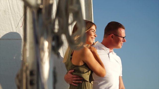 Owners of big yacht is boating in evening time. Man is embracing his beautiful wife, speaking something and smiling
