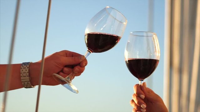 Two crystal glasses with wine on background of mast, white sail and blue sky. Man and woman are holding their and clinking by them, luxury celebration.
