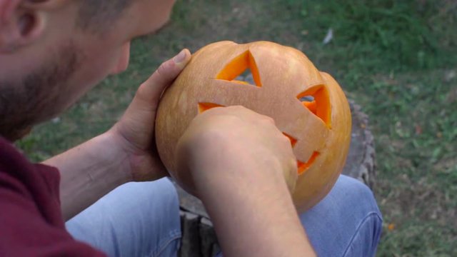 Man carves from a pumpkin Jack-o'-lantern in the backyard on a tree stump