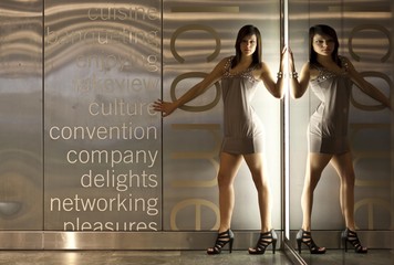 Young dark-haired woman wearing a beige dress and high heels posing in front of a wall with lighting