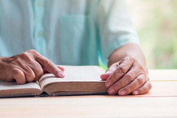 old man reading book on light brown wooden table surface, pointing with right index finger, selective focus