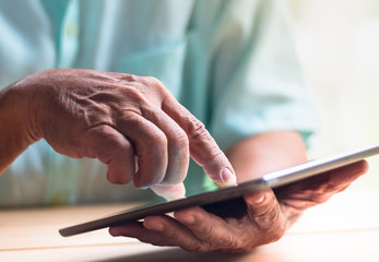 old man hold tablet with left hand and touch screen with right index finger on light brown wooden table surface