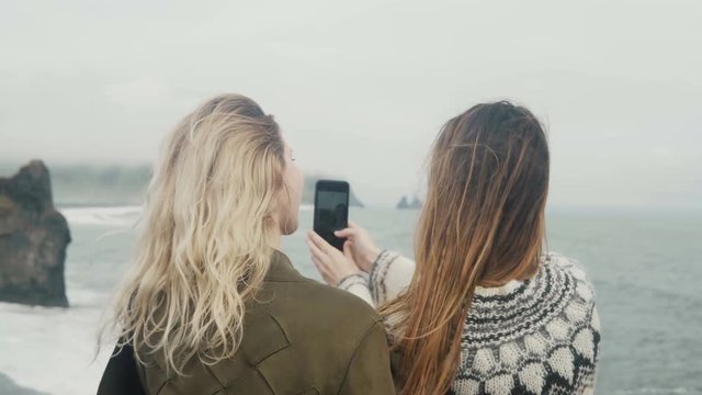 Two happy women taking selfie photos on smartphone, girls spending time on the beach near the troll toes in Iceland.