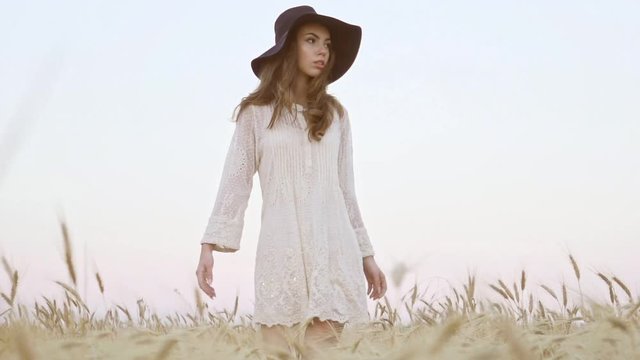View from below of young pretty woman in white dress and black hat walking in the field