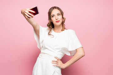 Portrait of a young attractive woman making selfie photo with smartphone on a pink background