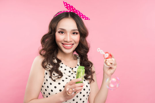 Portrait of a young woman playing with soap bubbles over pink background.
