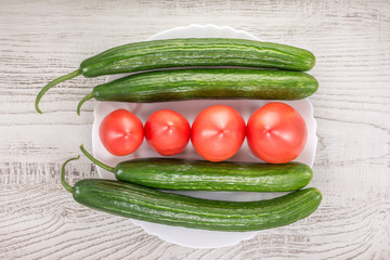 Four green cucumbers and four red tomatoes in a white plate on wooden background.Top view.
