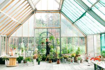 Young cute lady working at the greenhouse wearing linen dress and leather belt with florist tools. Bunch of wildflowers in her hands.