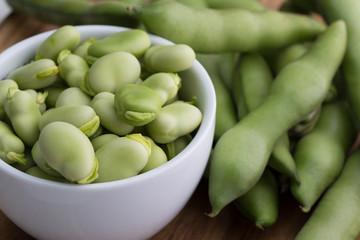 Close up fresh raw broad beans in a white bowl on a wooden cutting board surface