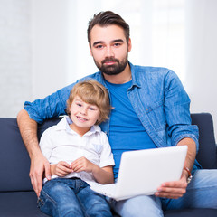 computer and internet concept - young father and little son sitting on sofa with laptop