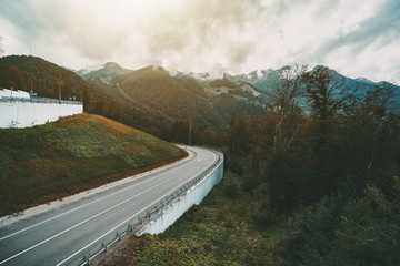 Beautiful autumn landscape with bending mountain highway bending after the hill, overcast sky, hills ridge in background overgrown with greenery and low rainy clouds, Russia, Estosadok district