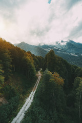 Wide-angle shooting from high above of dirt road stretching into distance between trees in mountains of Estosadok, Sochi district Russia; hills ridge in background with low clouds of overcast sky