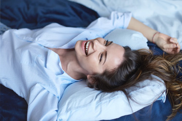 .Attractive cheerful brunette female stretching in bedroom while lying in bed wearing blue shirt and jeans, laughing out loud with charming smile, full of energy, feeling excited about new happy day.