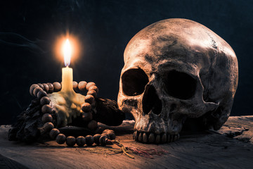 still life photography : human skull and light of burning candle with wood beads rosary on old wood against dark background
