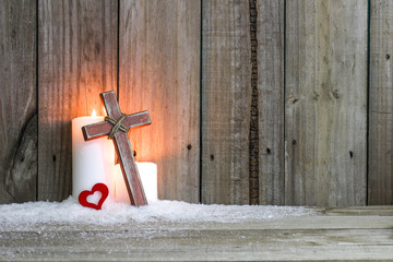 White holiday candles in snow by red heart and wooden cross with rustic wood background