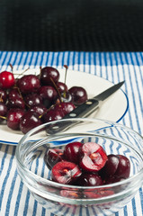  Preparing glass bowl of halved organic cherries, a cherry sauce, plate whole cherries with stems, round, white plat, blue rim, pairing knife, blue and white stripped table cloth