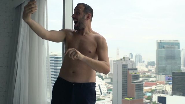 Topless man taking selfie photo with cellphone by window at home
