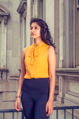 East Indian American college student with long curly hair studying in New York, wearing sleeveless orange shirt, striped pants, standing by vintage style office building. Instagram filtered effect..