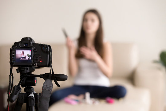 Young female videoblogger sitting on sofa, records commercial beauty product review for personal video blog. Vlogger uses contemporary photo camera to create footage for popular internet vlog at home.