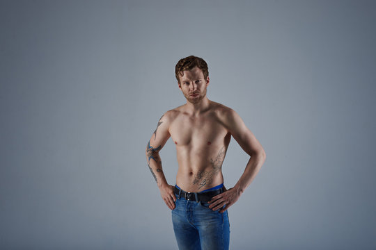 Horizontal shot of good looking bearded red haired guy with beautiful athletic body modeling in studio, posing shirtless, keeping hands on his waist and staring at camera with serious confident look