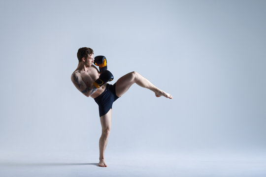 Isolated picture of young bearded European kickboxer standing barefooted on studio floor and kicking air in front of him, looking ahead with focused expression on his face. Martial arts concept