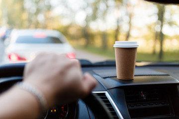 Fototapeta na wymiar Paper Cup with coffee on the dashboard of the car blurred green background. Without focus the man's hand holds the steering wheel