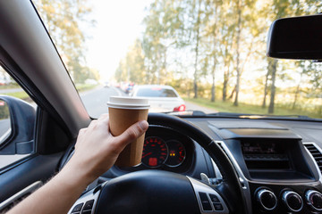Close up young man car driver drink coffee, hand holding a Paper Cup of coffee in the background steering the car dashboard blurry green background.