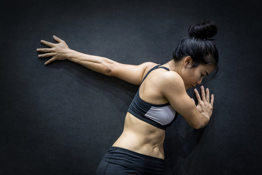 young asian woman wearing sportswear stretching her arm on black wall in fitness gym, exercise for healthy lifestyle concept