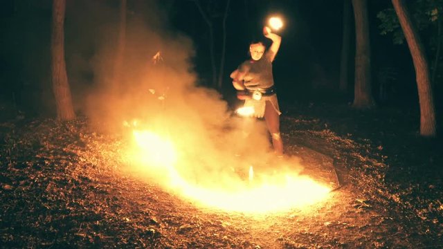 Fiery performance in the woods at night. The girl performs tricks with fire, the assistant in the robes helps her.