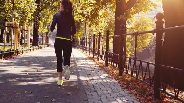 Runner Woman Running in the Sunny City Park Exercising Outdoors. Steadicam STABILIZED shot, SLOW MOTION 120 fps. Sportswoman Listening to Music during Morning Training. Healthy Lifestyle. Lens Flare.