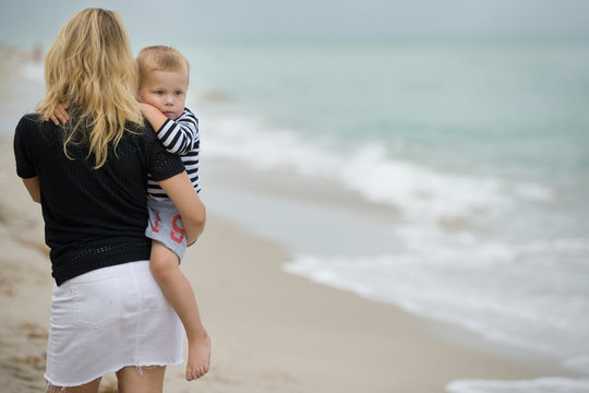 Portrait of cute toddler boy in the arms of his mother while walking along the beach. Family watching sea waves. Young blond woman holding child.
